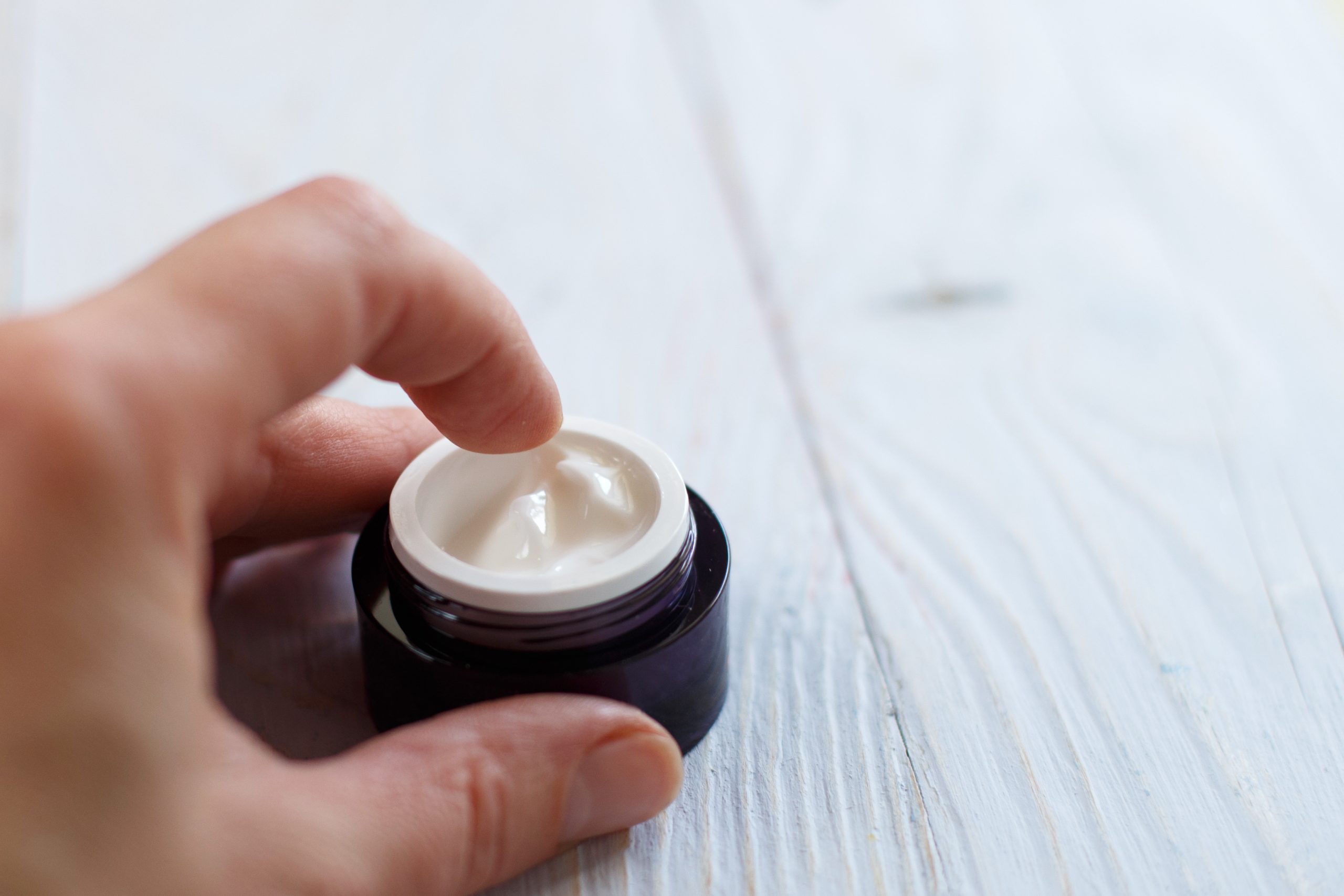 Hands of a woman with a jar of face cream close up applied to the finger of woman's hand on light blue wooden background