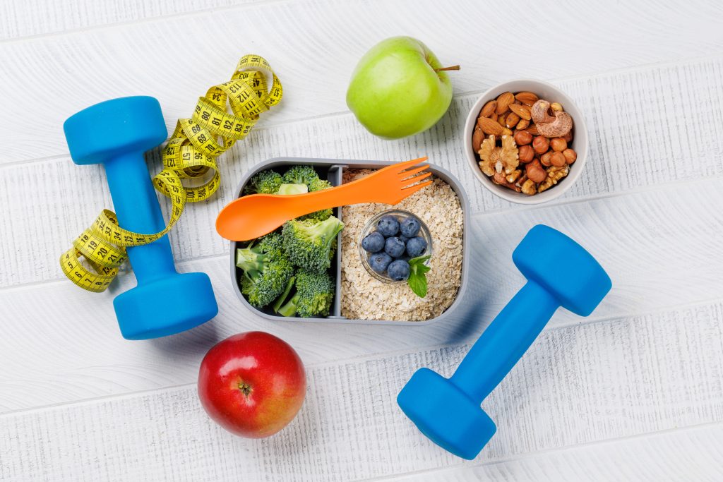 A healthy lunch of fruit and veggies with two dumb bells and a measuring tape.